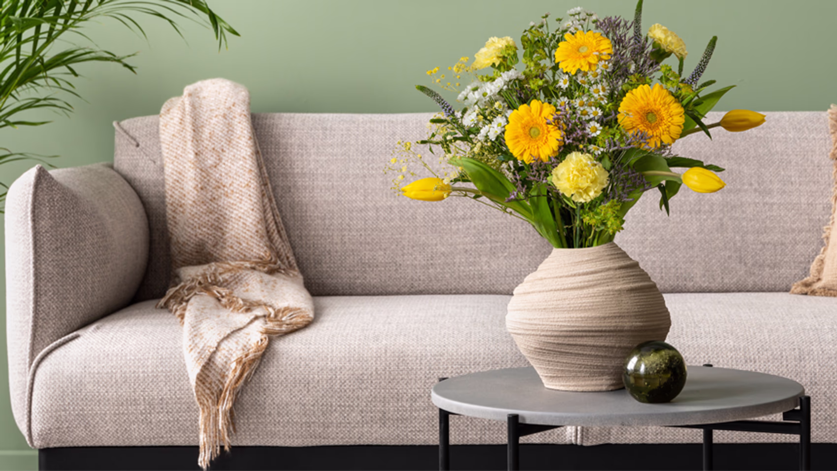 A landscape shot of a beige textured vase on a grey coffee table filled with a mix of yellow and white flowers surrounded by green foliage. The background features a beige sofa and a sage green wall.
