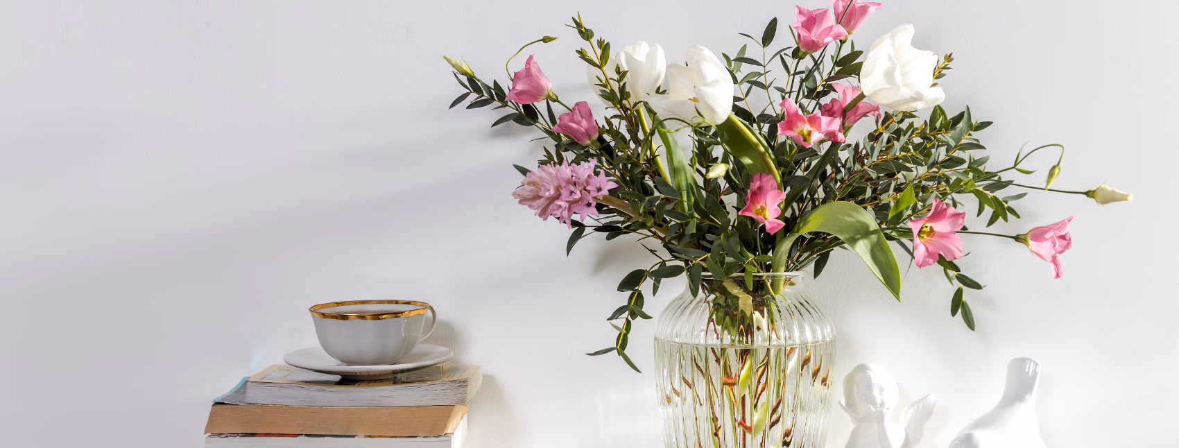 A bouquet of pink and white flowers in a clear ribbed glass vase on a shelf next to a stack of books with a teacup on the top.