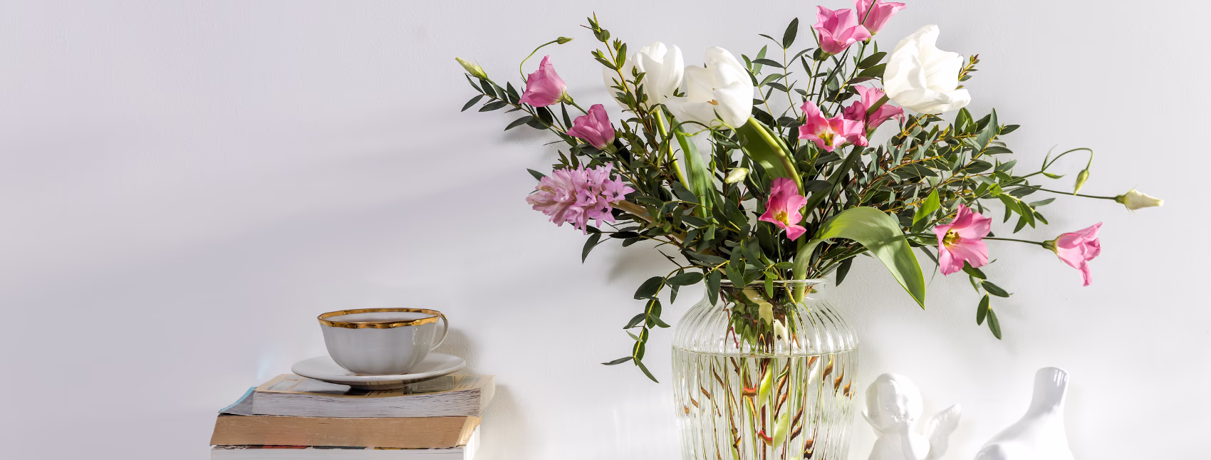 A bouquet of pink and white flowers in a clear ribbed glass vase on a shelf next to a stack of books with a teacup on the top.