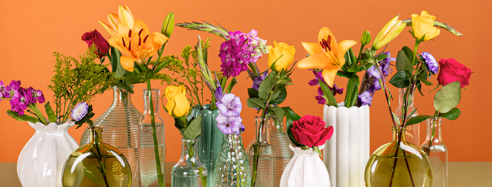 A landscape shot of several different vases in clear, green, and white with different stems in each from orange lilies to dark pink and yellow roses and greenery