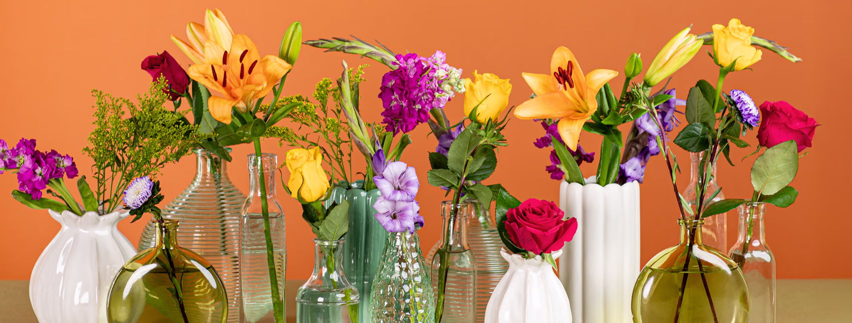 A landscape shot of several different vases in clear, green, and white with different stems in each from orange lilies to dark pink and yellow roses and greenery